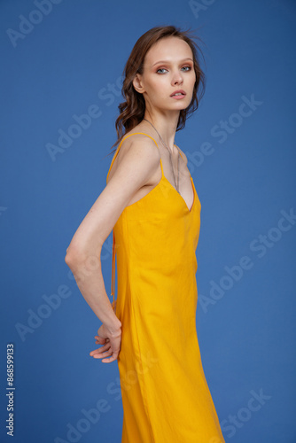 High fashion photo of a beautiful skinny young woman in pretty long yellow dress posing on blue background. Slim figure.