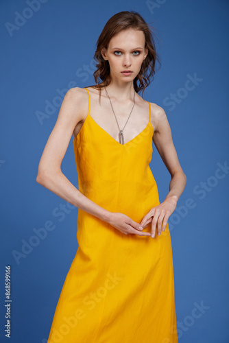 High fashion photo of a beautiful skinny young woman in pretty long yellow dress posing on blue background. Slim figure.