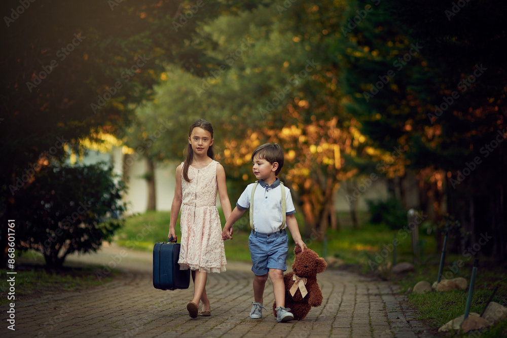 Holding hands, suitcase and teddy bear with children walking on path in forest for travel to school. Destination, evening or luggage with lost brother and sister kids outdoor in park or woods Holding hands, suitcase and teddy bear with children walking on path in forest for travel to school. Destination, evening or luggage with lost brother and sister kids outdoor in park or woods