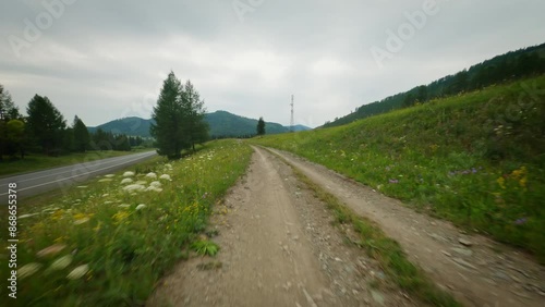 A FPV copter flies over a dirt road in the mountains. Two highways are asphalt and dirt, cloudy weather and mountains with green trees.
