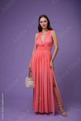 High fashion photo of a beautiful elegant young woman in pretty long coral red dress, cream handbag posing on purple, lilac background. Slim figure. 