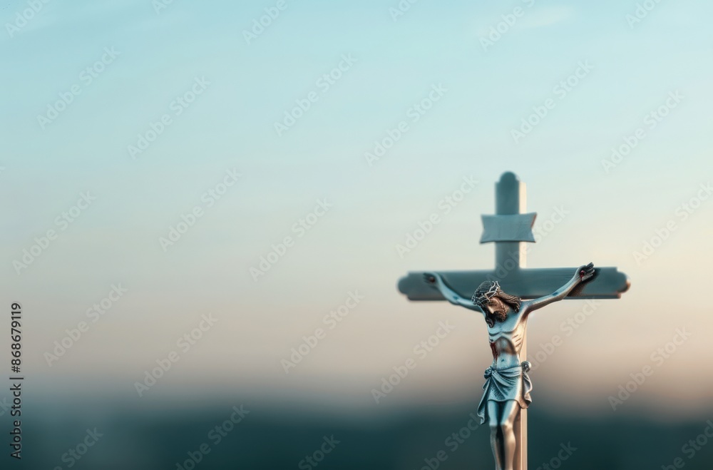 A silver crucifix stands against a soft, blue sky with a subtle pink hue. The figure of Jesus is in focus, while the background is blurred.