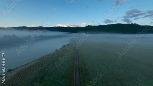 The FPV copter flies low to the ground. Shooting early in the morning at dawn in a thick fog. The copter flies low above the ground, past the trees. Mountains in the background, rivers, trees.