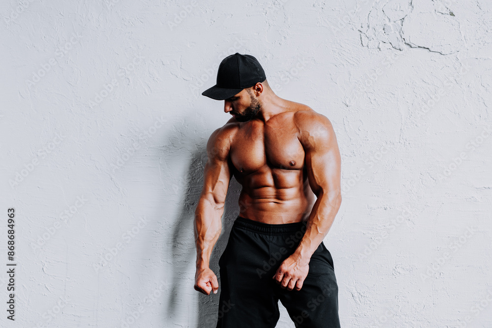 Naklejka premium Young strong man bodybuilder in cap on white wall background
