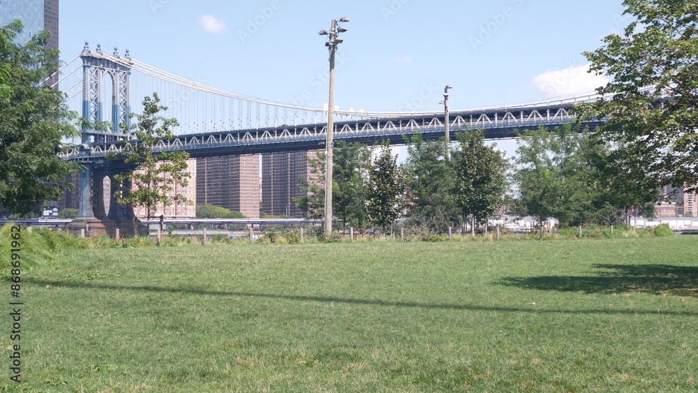 Manhattan Bridge to Brooklyn Dumbo. New York City symbol, USA travel destination. Architecture of United States, tourist landmark. Cable-stayed bridge. Park greenery, green grass picnic lawn by river. Manhattan Bridge to Brooklyn Dumbo. New York City symbol, USA travel destination. Architecture of United States, tourist landmark. Cable-stayed bridge. Park greenery, green grass picnic lawn by river.