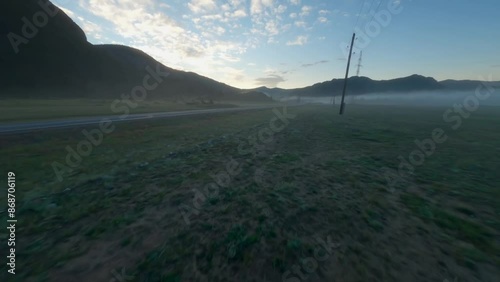 The FPV copter flies low to the ground. Shooting early in the morning at dawn in a thick fog. The copter flies low above the ground, past the trees. Mountains in the background, rivers, trees.