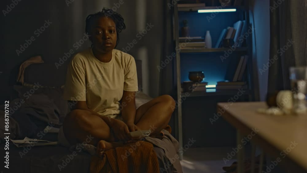 Full portrait of exhausted African American woman with swollen eyes sitting in corner of bed with crossed legs and looking at camera, having sleep disorder due to ptsd