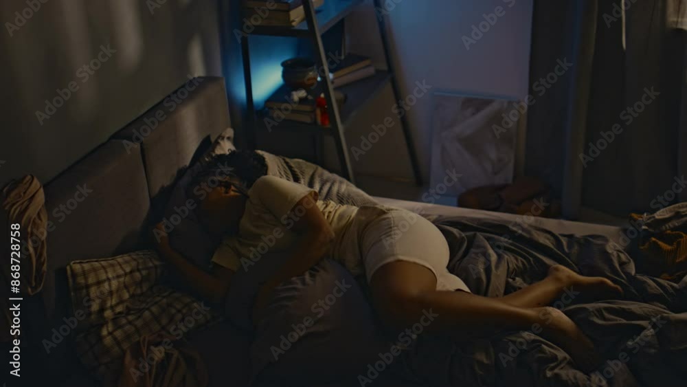 High angle side shot of young Black woman with ptsd rolling over in bed, waking up from terrifying nightmares and sitting up at night, living with traumatic memories