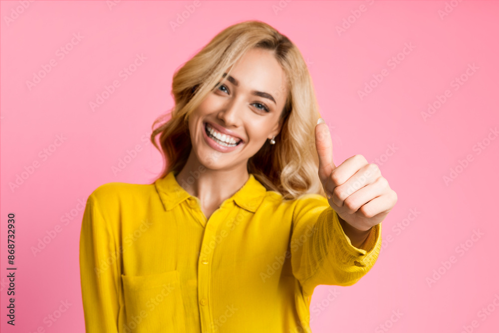 A young woman with long blonde hair is smiling and giving a thumbs up gesture. She is wearing a bright yellow button-down shirt and the background is a solid pink. A young woman with long blonde hair is smiling and giving a thumbs up gesture. She is wearing a bright yellow button-down shirt and the background is a solid pink.