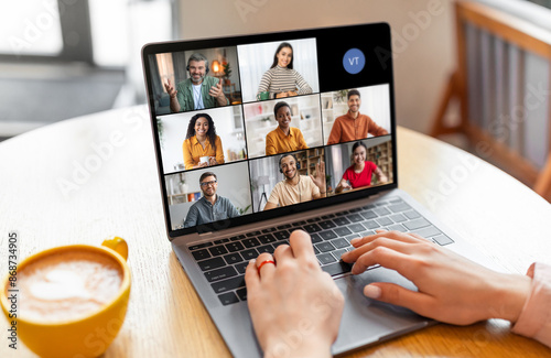 Lady sits at a table in a coffee shop, using a laptop to participate in a video conference with a group of people. The screen shows six video tiles of the participants photo