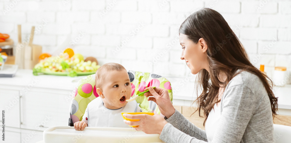 A young mother is feeding her baby in a high chair in the kitchen. The baby is sitting in a brightly colored high chair and is looking at the mother with a wide-eyed expression, copy space A young mother is feeding her baby in a high chair in the kitchen. The baby is sitting in a brightly colored high chair and is looking at the mother with a wide-eyed expression, copy space