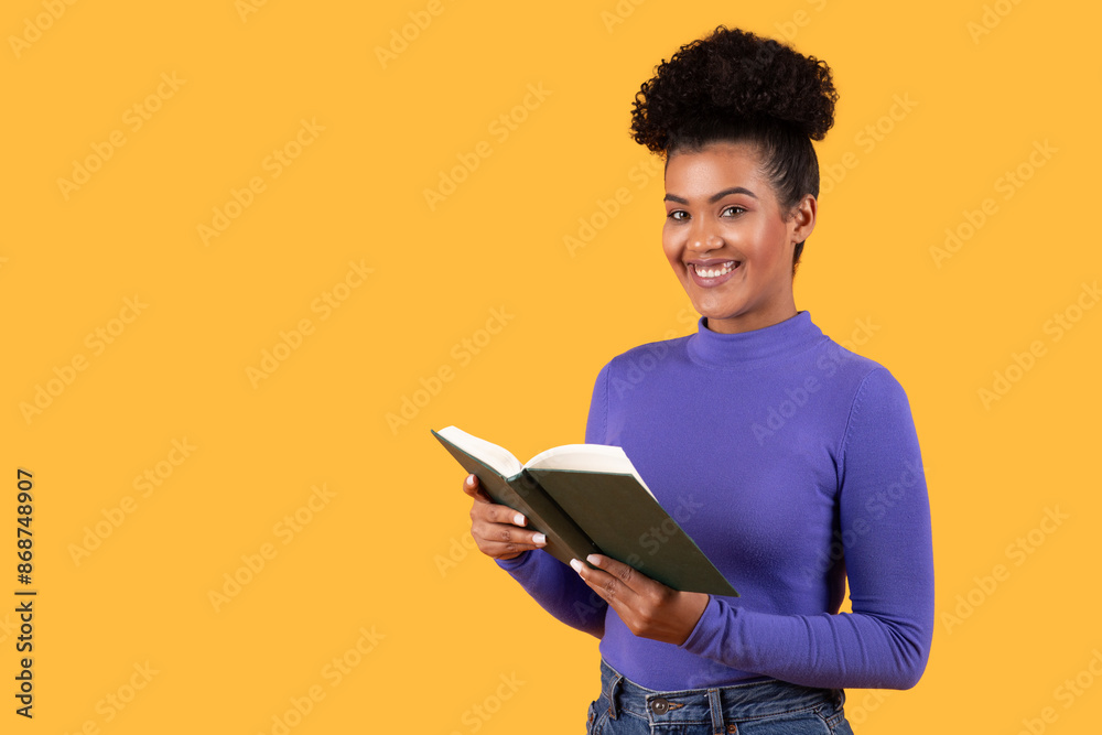 This image shows a young woman with dark skin and curly hair, smiling while holding a book open in front of her. She is wearing a purple shirt and jeans and is standing in front of yellow background. This image shows a young woman with dark skin and curly hair, smiling while holding a book open in front of her. She is wearing a purple shirt and jeans and is standing in front of yellow background.