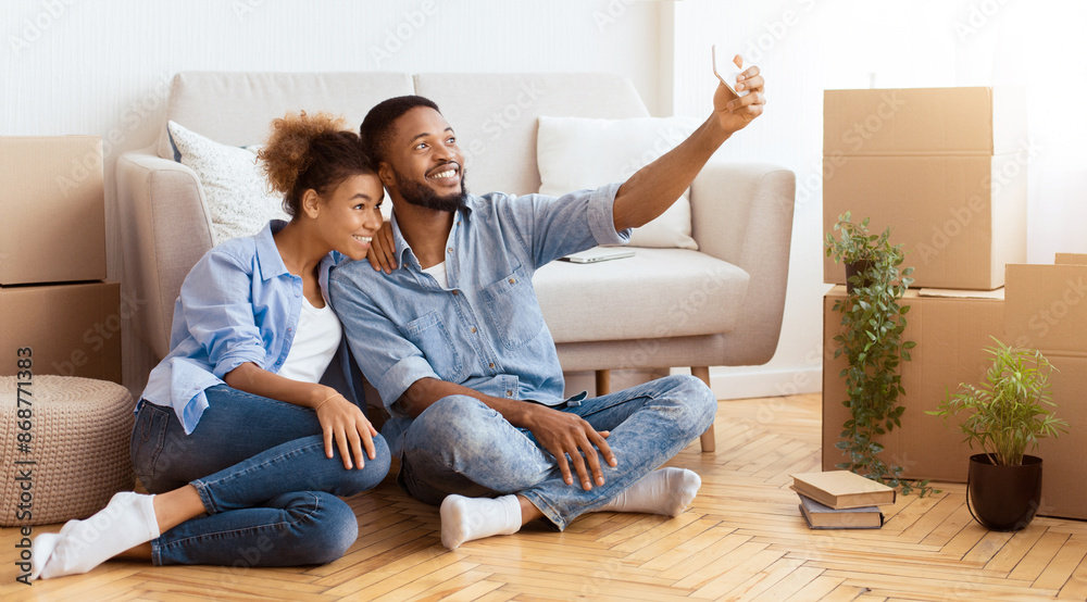 Fototapeta premium Moving New Home. African American Spouses Making Selfie On Smartphone Sitting On Floor Indoor. Panorama, Empty Space