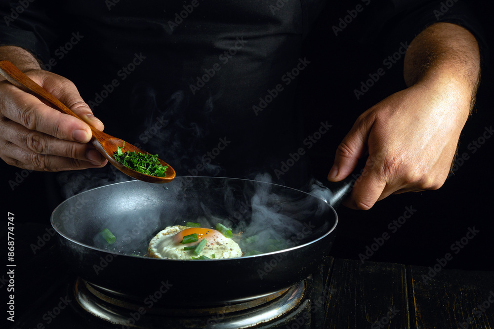 Fototapeta premium The chef cooks a delicious fried eggs in a frying pan. Adding herbs with a spoon to the hot fried egg.