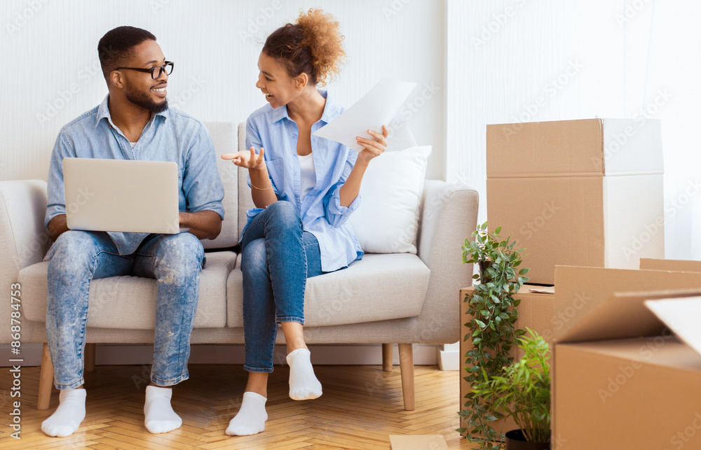 Buying Own House. Happy Afro Couple Using Laptop Reading Ownership Papers Sitting On Couch After Moving Apartment. Copy Space Buying Own House. Happy Afro Couple Using Laptop Reading Ownership Papers Sitting On Couch After Moving Apartment. Copy Space