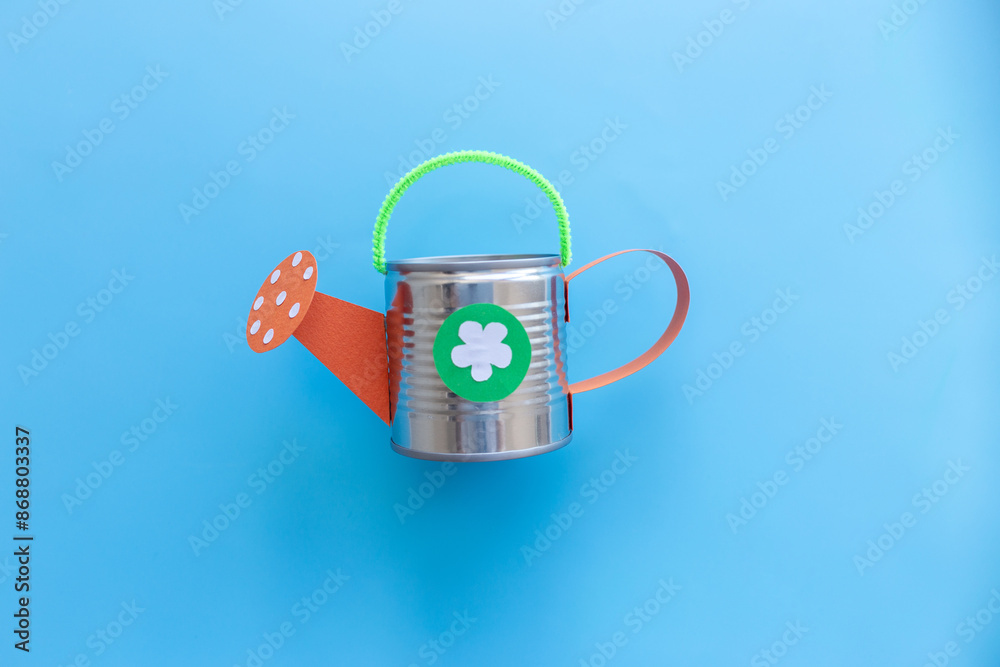 Obraz premium crafted watering can with green handle, orange spout, and recycled tin can body. Solid light blue backdrop provides contrast, suitable for discussions on recycling, gardening, crafts, DIY endeavors