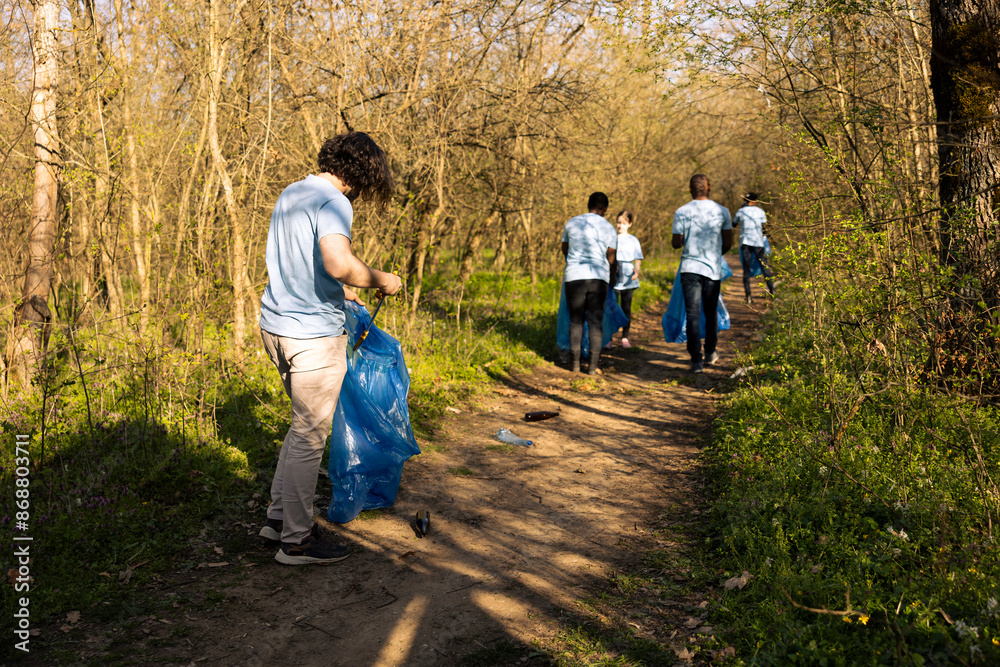 Fototapeta premium Male environmentalist collecting trash with a long claw tool and bag, grabbing plastic waste and rubbish to tidy up the forest area. Young man cleaning the natural habitat, global pollution.