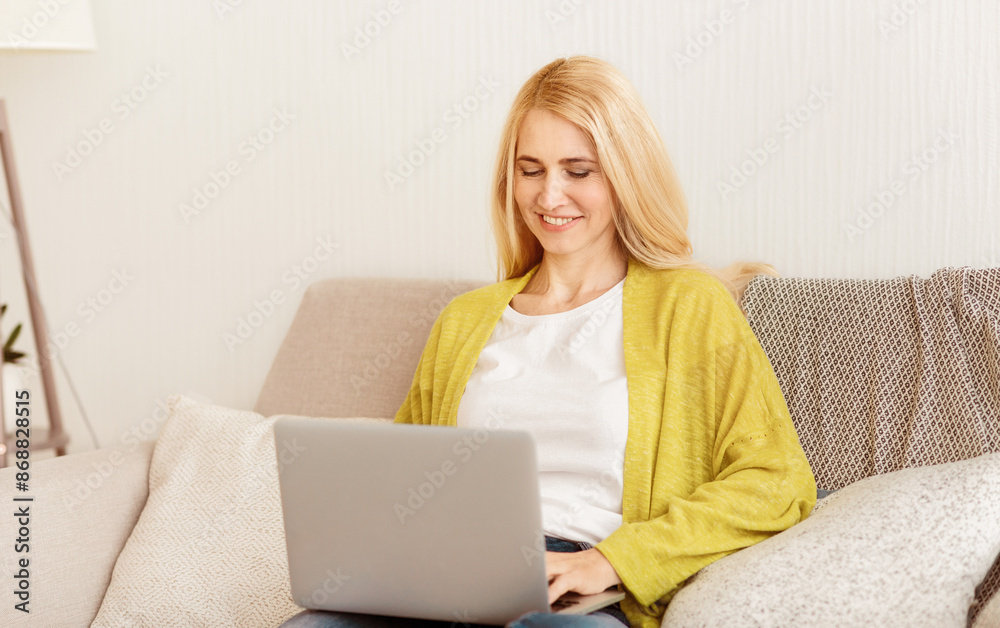 A woman in a yellow cardigan sits on a sofa and uses a laptop. A woman in a yellow cardigan sits on a sofa and uses a laptop.