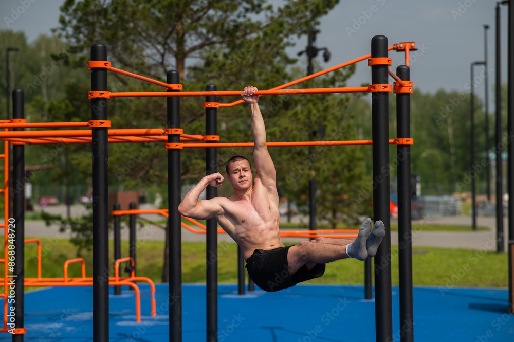 Fototapeta premium Young muscular man hanging by one hand on a horizontal bar outdoors. 