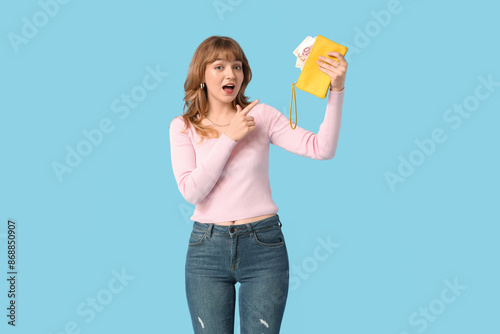 Beautiful young woman pointing at yellow wallet with money on blue background