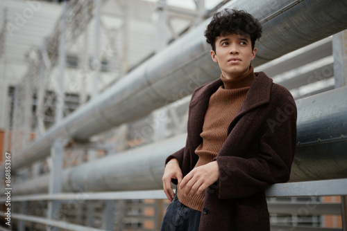 Fashionable young man in brown sweater and black pants posing in front of industrial building with pipes