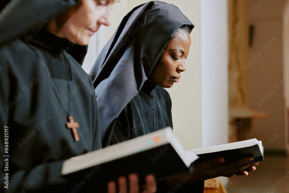 Selective focus medium closeup of two ethnically diverse nuns holding books in hands and reading religious texts Selective focus medium closeup of two ethnically diverse nuns holding books in hands and reading religious texts