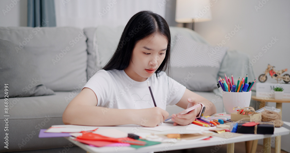 Portrait of Young Asian woman sitting on the floor uses a mobile phone to search for ideas for greeting cards. Holiday hobbies, crafts, DIY cards Portrait of Young Asian woman sitting on the floor uses a mobile phone to search for ideas for greeting cards. Holiday hobbies, crafts, DIY cards
