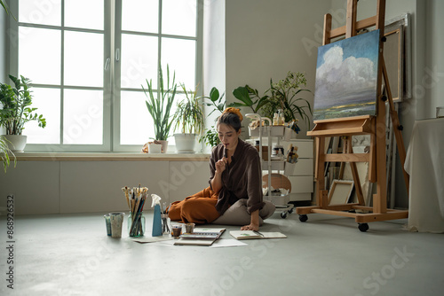 Focused female artist sitting on floor, looking at sketches, drawings in sketchbook with interest. Thoughtful inspired woman immersed in creative process. Girl in home art studio among oil paintings.