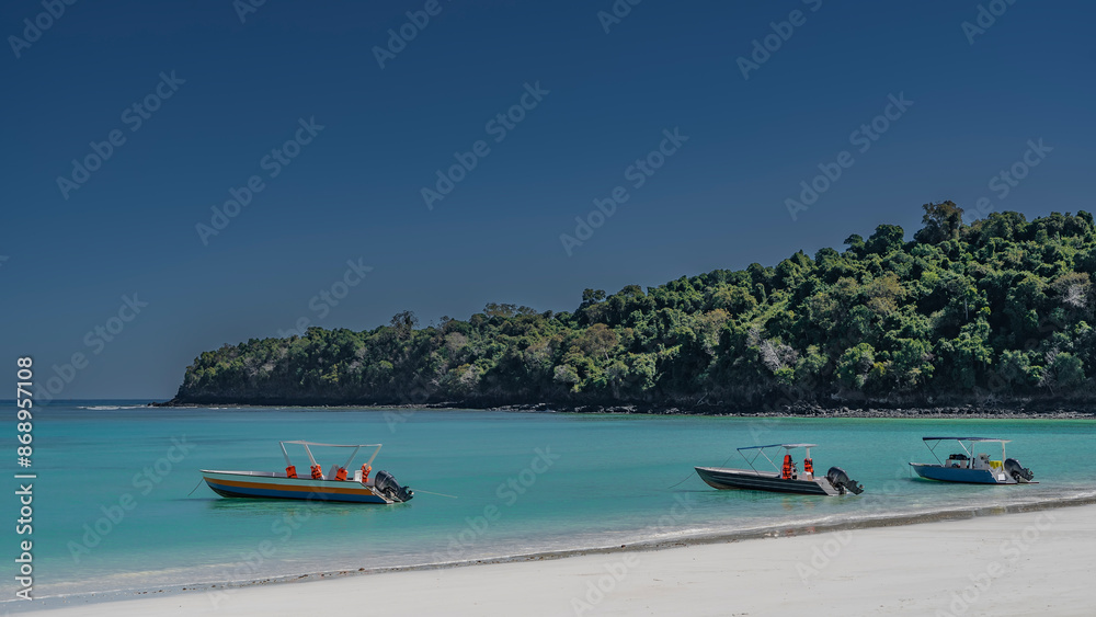 Motor boats are anchored near the shore. The aquamarine ocean is calm. A clean white sandy beach. A green hill against a blue sky. Madagascar.