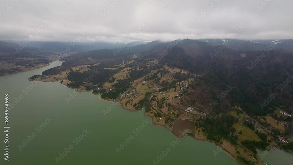 Aerial view of wonderful Colibita lake along forested hills with scattered rural homes on a cloudy day in Transilvania, Romania. Beautiful green landscape with Carpathian mountains in background. Aerial view of wonderful Colibita lake along forested hills with scattered rural homes on a cloudy day in Transilvania, Romania. Beautiful green landscape with Carpathian mountains in background.