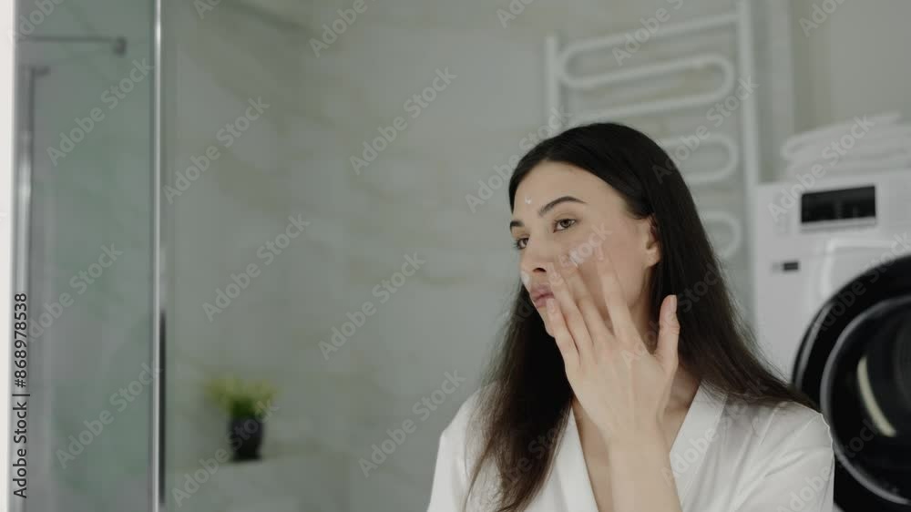 A woman in a modern bathroom applies skincare cream to her face, enhancing her healthy and glowing skin. The scene conveys self-care, beauty, and a daily skincare routine.Woman Applying Skincare Cream A woman in a modern bathroom applies skincare cream to her face, enhancing her healthy and glowing skin. The scene conveys self-care, beauty, and a daily skincare routine.Woman Applying Skincare Cream