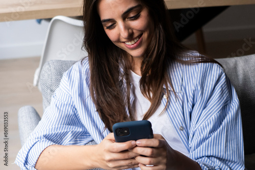 Happy pleasant millennial woman relaxing on comfortable couch, holding smartphone in hands. Smiling young lady chatting in social networks, watching funny videos, using mobile applications at home. photo