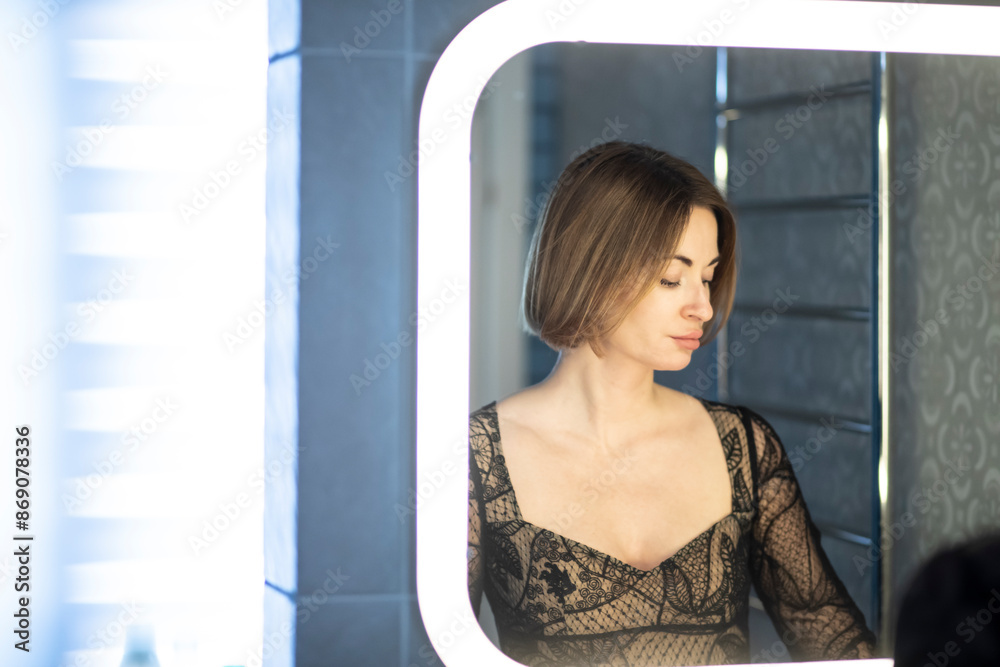 Fototapeta premium Fashionable beautiful young woman in stylish vintage clothing painting lips with a red lipstick in front of the mirror in lavatory at the night time. Attractive girl preparing for dating and party