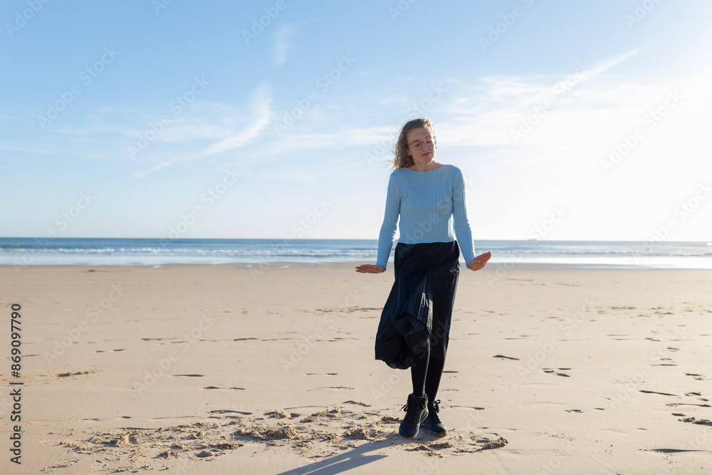 A woman is standing on a beach, wearing a blue shirt and black pants A woman is standing on a beach, wearing a blue shirt and black pants