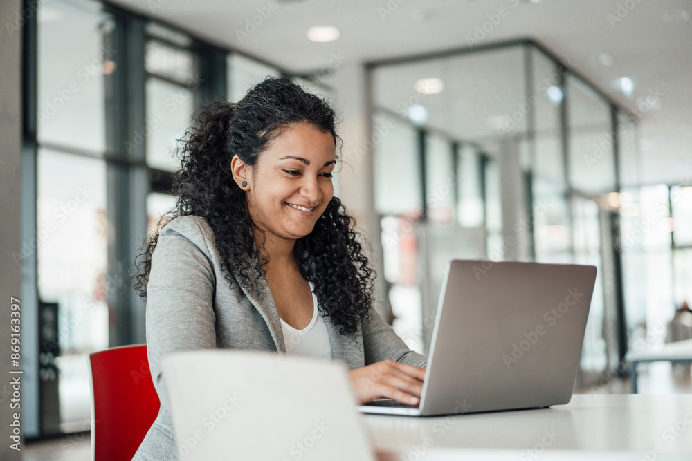 Smiling businesswoman using laptop and sitting at desk in office Smiling businesswoman using laptop and sitting at desk in office