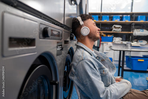 Young man listening to music through wireless headphones leaning on washing machine in laundromat