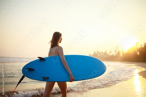 A young surfer woman walks by the ocean with a blue surfboard before surfing at sunset