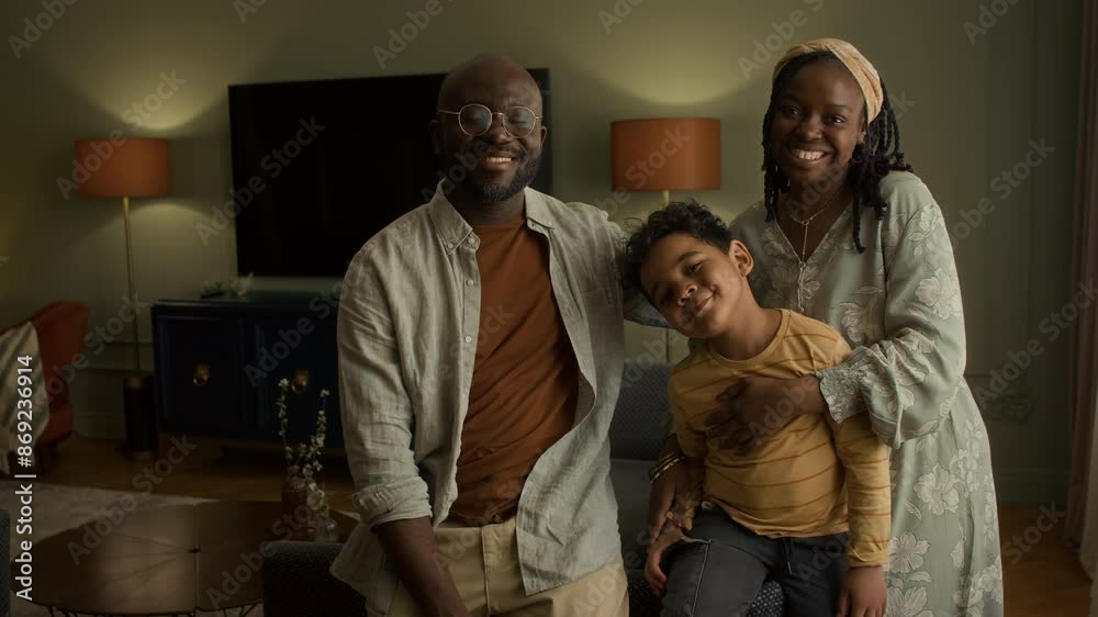 Medium portrait of smiling young middle class African American couple with 6-year-old son posing in living room in cozy home, mother hugging boy, looking at camera and smiling