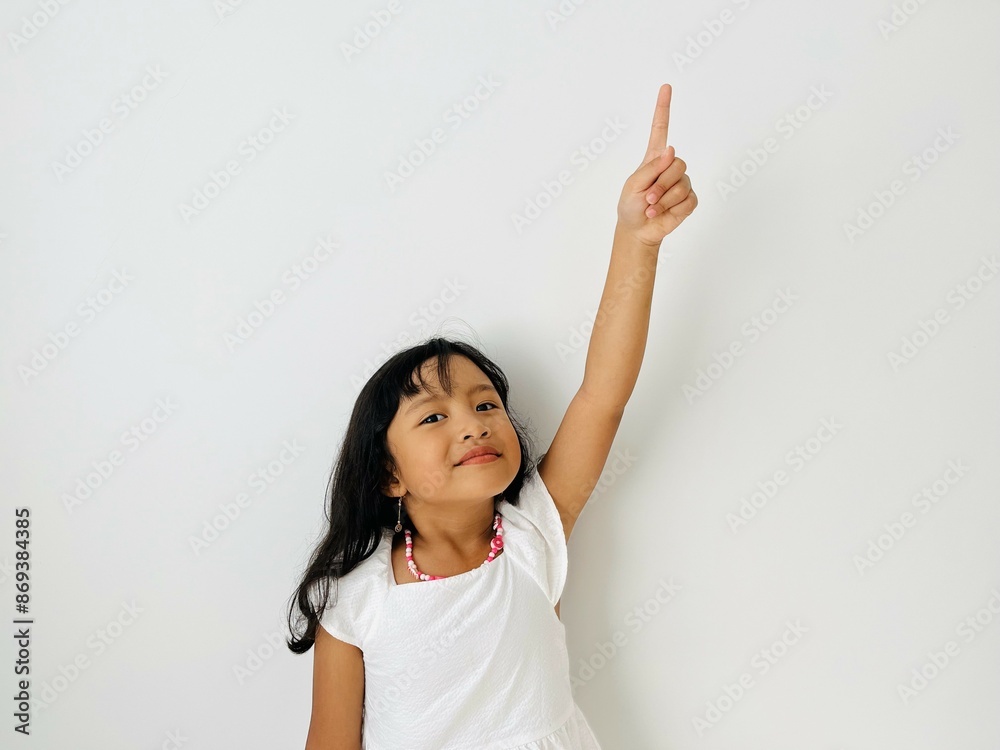 The little Asian girl raises her hand, points up with her index finger and smiles. A child in white dress on a white isolated background The little Asian girl raises her hand, points up with her index finger and smiles. A child in white dress on a white isolated background