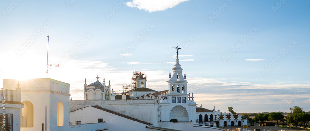 Fototapeta premium Sunlight bathes a traditional white building adorned with ornate blue tiles and a classic weather vane, set against a soft blue sky with wisps of clouds in the tranquil early morning