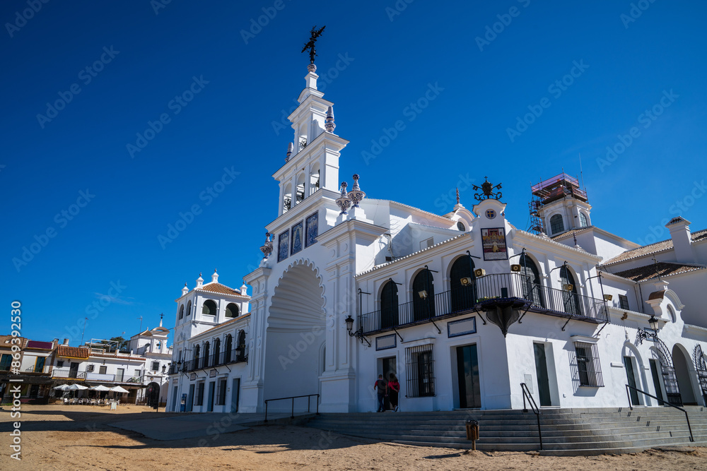 Obraz premium The pristine facade of the Hermitage of El Rocío gleams under the Spanish sun, with its elegant white walls and ornate religious detailing standing out against the clear blue sky.