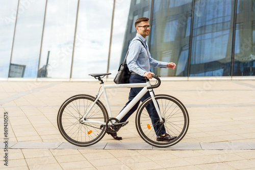 Smiling businessman walking his white bicycle, dressed in smart attire, in front of a modern glass building.