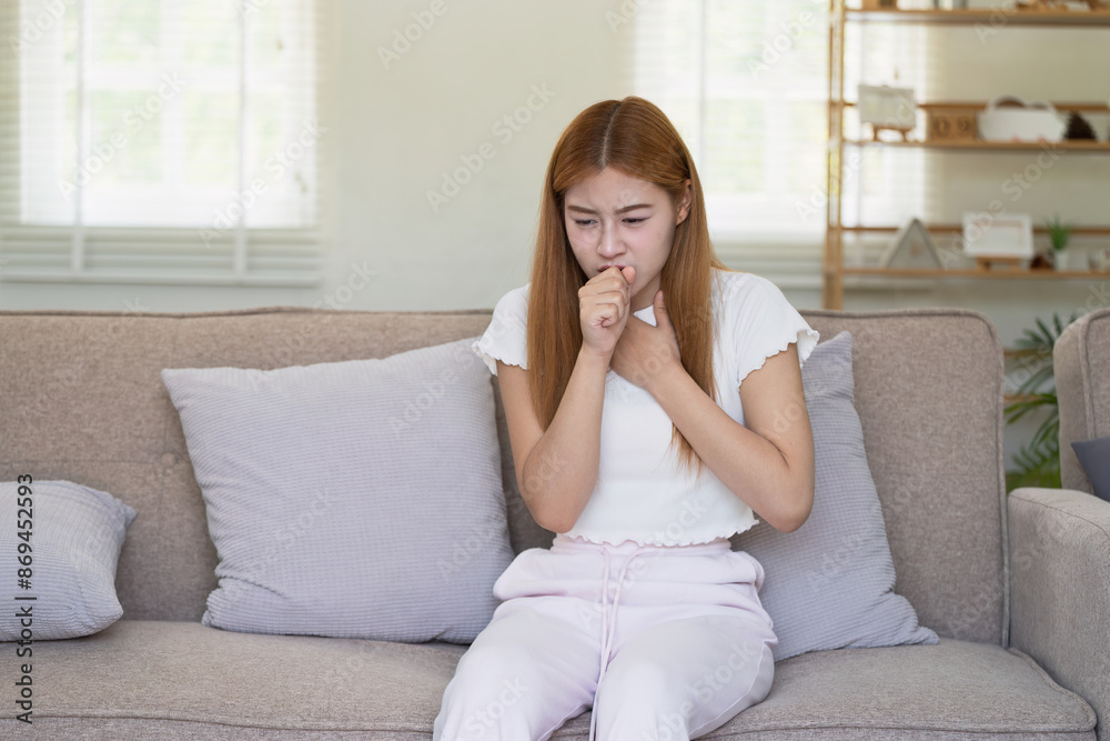 Young Woman Feeling Unwell and Coughing While Sitting on a Sofa at Home, Representing Illness and Sickness in a Domestic Setting Young Woman Feeling Unwell and Coughing While Sitting on a Sofa at Home, Representing Illness and Sickness in a Domestic Setting