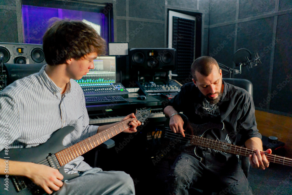 Obraz premium in the equipment of the recording room handsome man with a beard and tattoos and his young friend play the guitar in pairs and act out a song