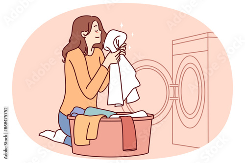 Housewife woman sits near washing machine and inhales fragrant smell freshly washed towel after using good laundry detergent. Girl takes out washed clothes from washing machine putting them in bowl