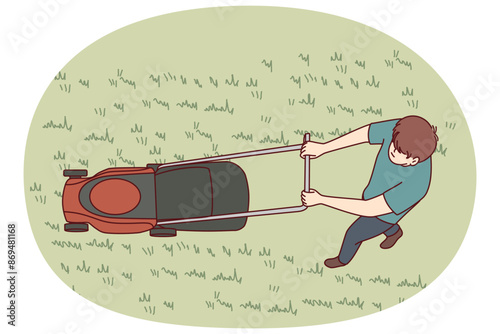 Man uses lawn mower to cut grass and wild weeds on golf course in park or in own backyard. Top view of young guy dressed in casual clothes with lawn mower working as gardener or janitor.
