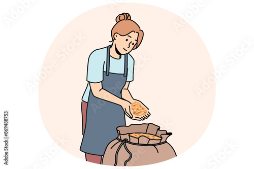 Woman stands near bag of malt for production of craft beer and collects hop grains in palms. Girl in apron works in agricultural factory preparing grains of prosso or wheat for processing.