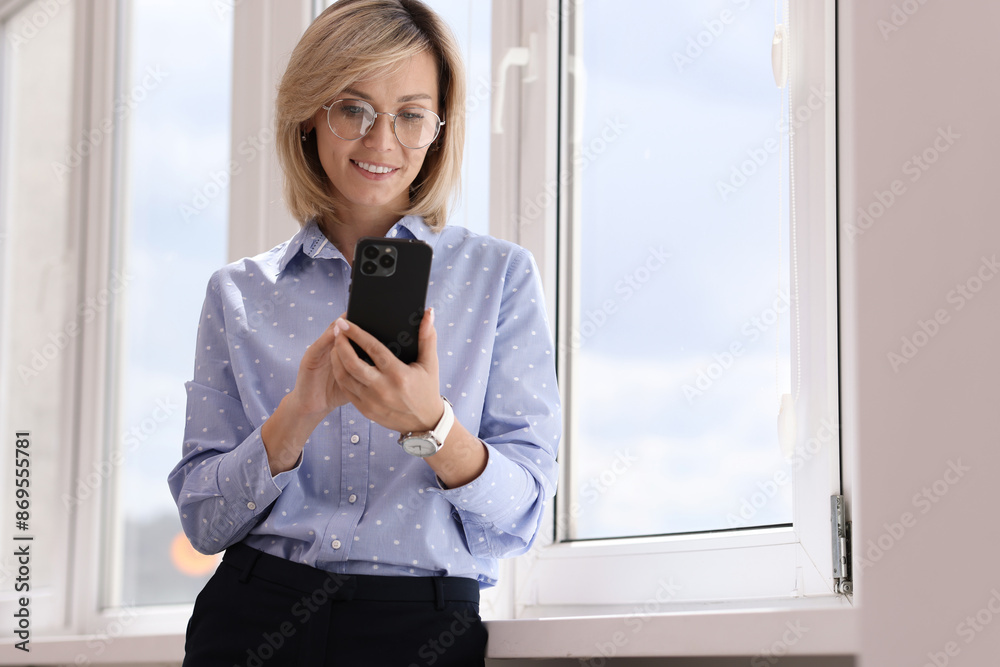 Fototapeta premium Happy woman using mobile phone near window indoors, space for text