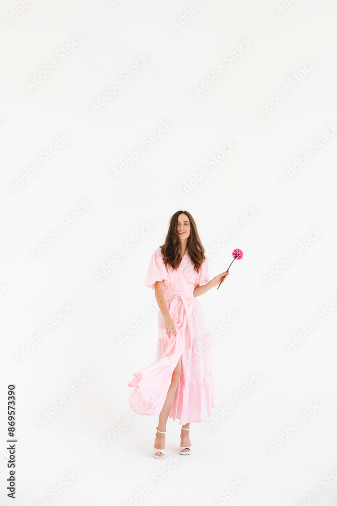 Model wearing casual outfit. Look in trendy minimalistic style. Pink dress with slit, belt, puff sleeves. Sexy sensual happy woman. Positive woman turn around and hold pink flower on white background. Model wearing casual outfit. Look in trendy minimalistic style. Pink dress with slit, belt, puff sleeves. Sexy sensual happy woman. Positive woman turn around and hold pink flower on white background.