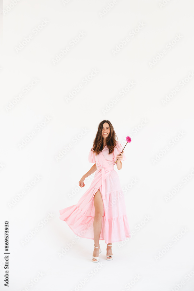 Model wearing casual outfit. Look in trendy minimalistic style. Pink dress with slit, belt, puff sleeves. Sexy sensual happy woman. Positive woman turn around and hold pink flower on white background. Model wearing casual outfit. Look in trendy minimalistic style. Pink dress with slit, belt, puff sleeves. Sexy sensual happy woman. Positive woman turn around and hold pink flower on white background.